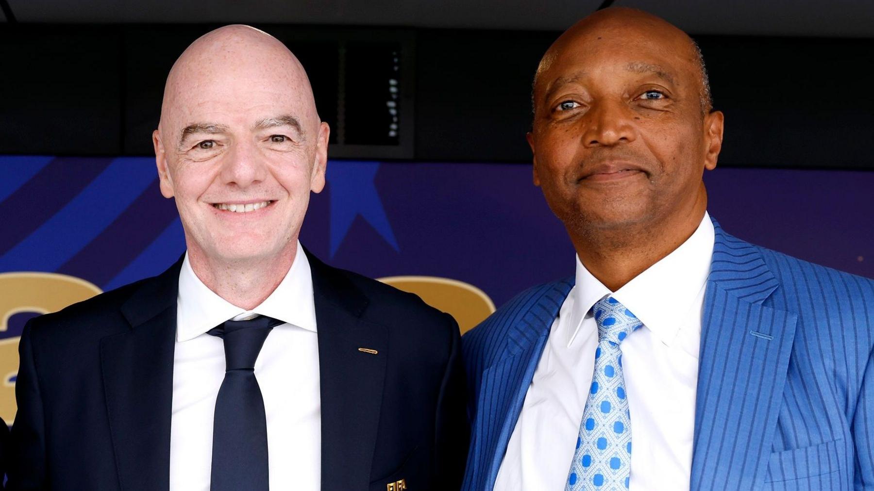 Gianni Infantino, wearing a dark blue suit jacket, white shirt and dark blue tie, stands next to Patrice Motsepe, who wears a light blue pinstriped suit jacket, white shirt and patterned blue and silver tie. Both men are seen from chest up as they smile into the camera