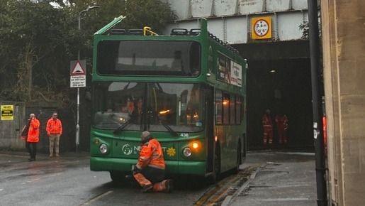 Green double decker school bus pictured in the middle of the road. Three men can be seen in orange high vis stood around the bus. The railway bridge can be seen behind the men.