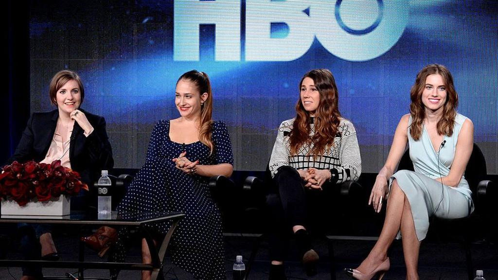 Four actresses sit in a line on black chairs while on stage with the lettering HBO seen in the background.