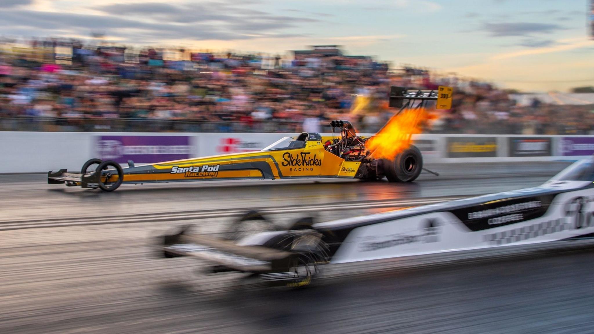 Susanne Callin racing in her yellow Top Fuel dragster at Santa Pod, near Northampton UK, in 2025. Tongues of orange flame are coming out of the back of the car as it speeds along the drag strip. In the foreground, the front part of the black and white dragster she is racing against can be seen, it is blurred because the camera is focused on Callin's car and the speeds are very high.