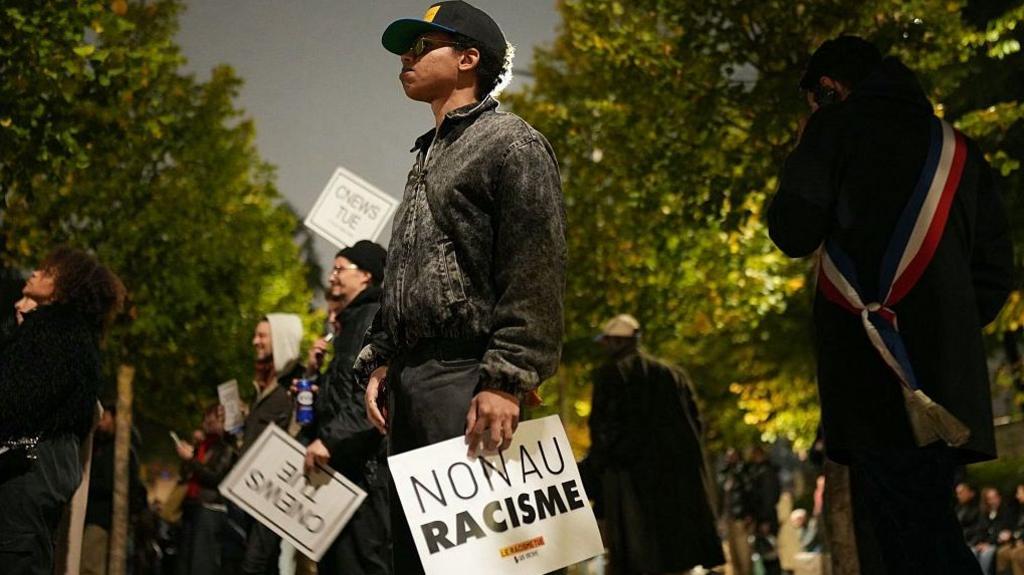 A man stands solemnly in a line of people holding anti-racism signs. His sign says 'non au racisme'
