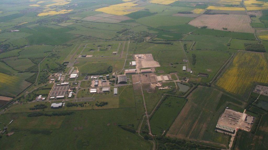 An aerial view of green fields and large warehouse buildings or plane hangars.