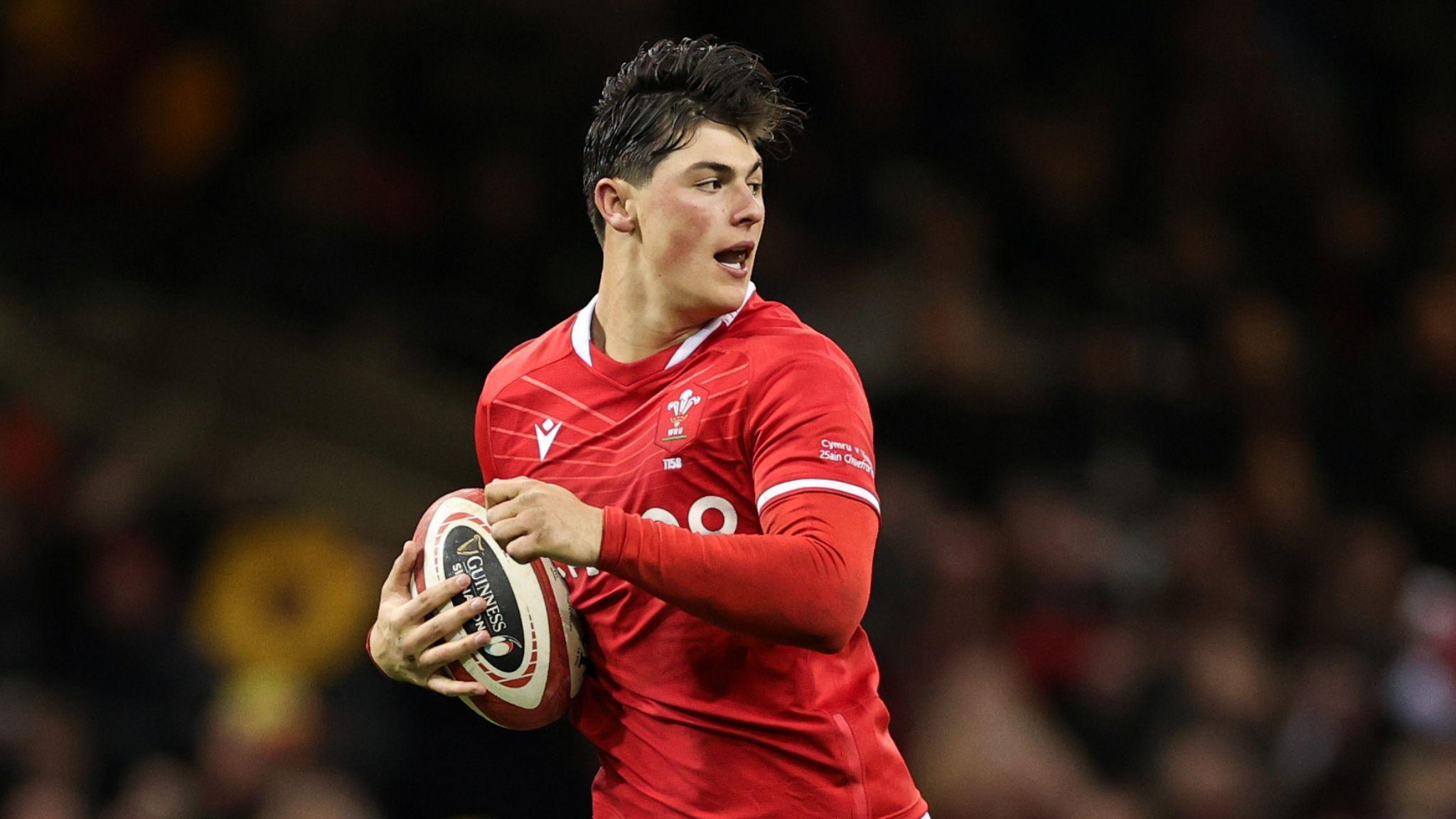 Louis Rees-Zammit looks behind him as he runs with the ball under his right arm in a red jersey of Wales.