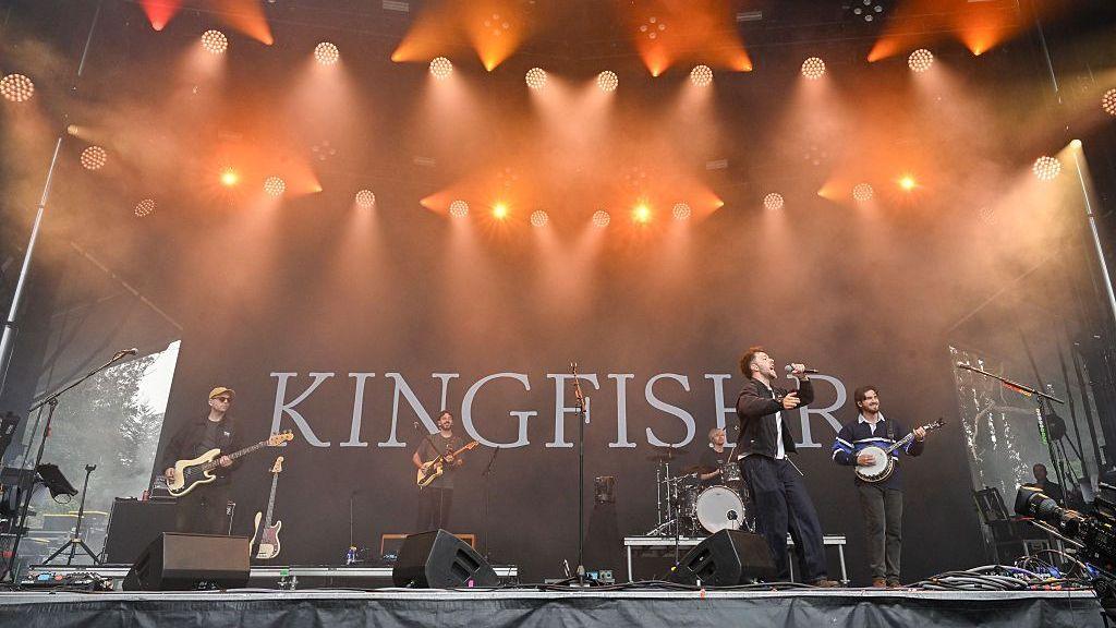 A wide shot of five men with guitars, performing on a lit up outdoor stage. The logo KINGFISHR is projected behind them.