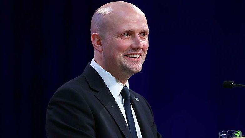 a bald man in a dark jacket, white shirt and dark tie is smiling as he gives a speech