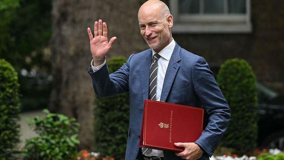 Stephen Kinnock arrives in Downing Street carrying a red Welsh secretary file and raising a hand in greeting. Trees, trimmed shrubs and flowerbeds can be seen in the background.