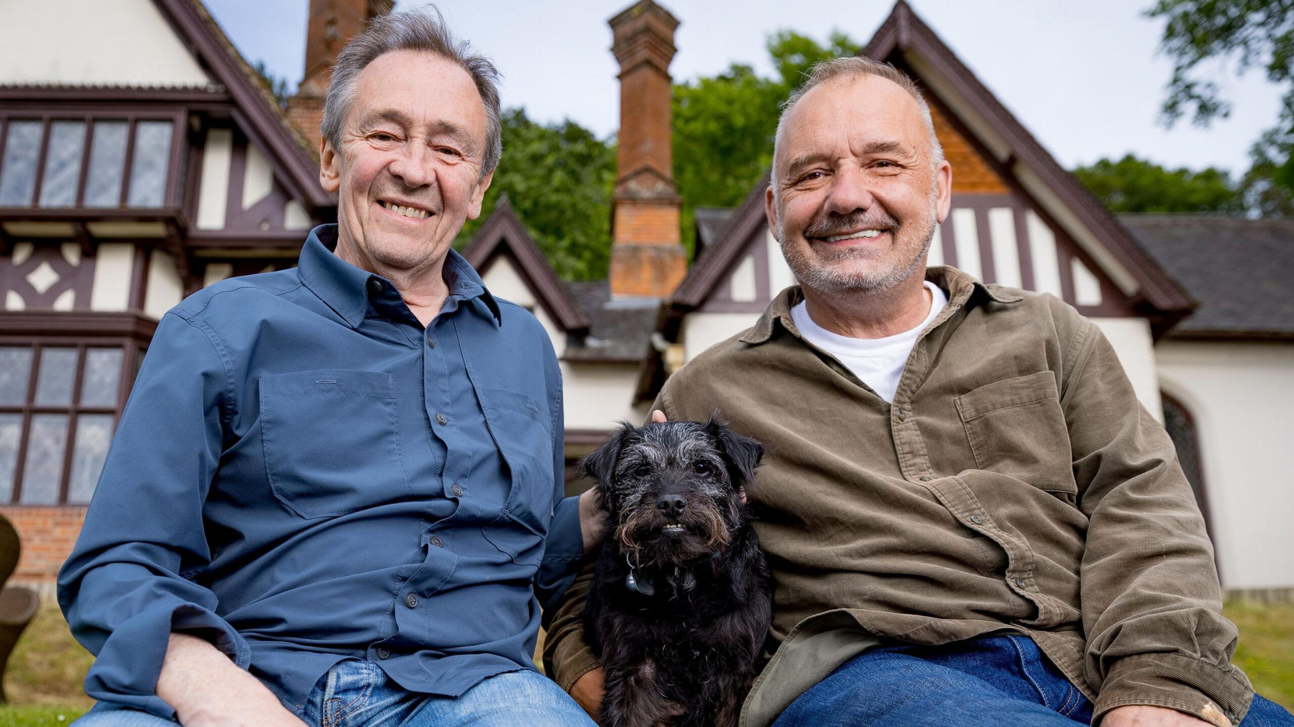 Bob Mortimer and Paul Whitehouse pose for a photo in front of a white house with black dog Ted in the middle.