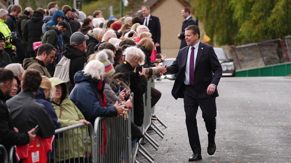 Rhun ap Iorwerth is walking on a road and shaking hands of many people who are stood behind a metal barrier.