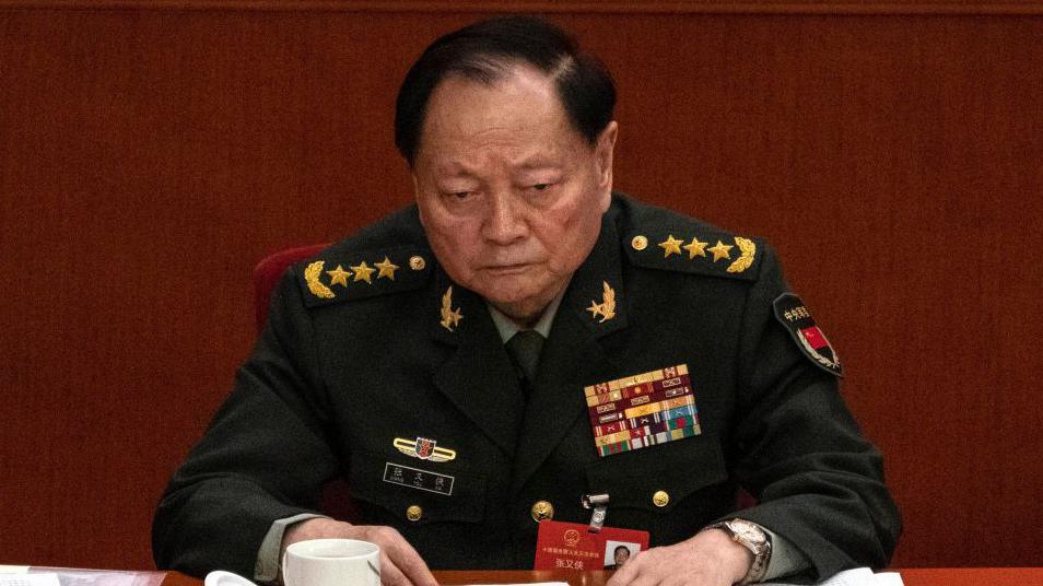 Zhang Youxia sits at table wearing dress military uniform adorned with medals and three golden stars and golden wings on epaulettes.