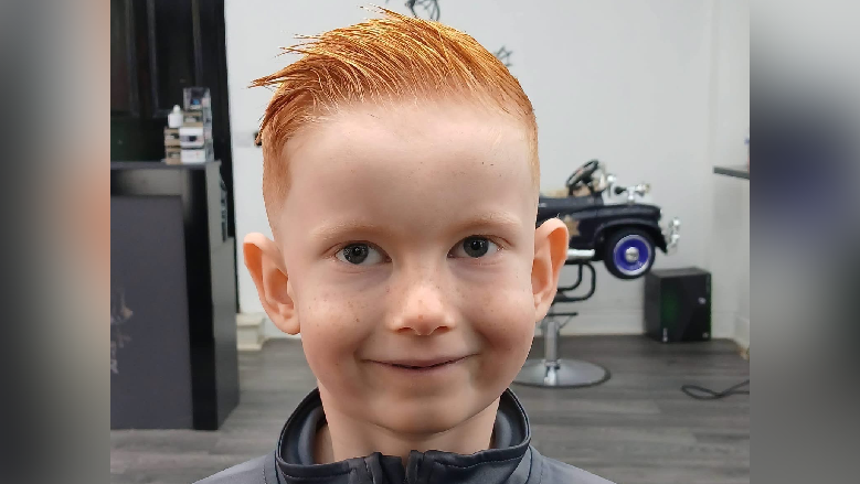 Harrison pictured at hairdressers smiling. He wears a grey zip top and has short red hair styled with gel and a small toy car child's seat is visible behind him.