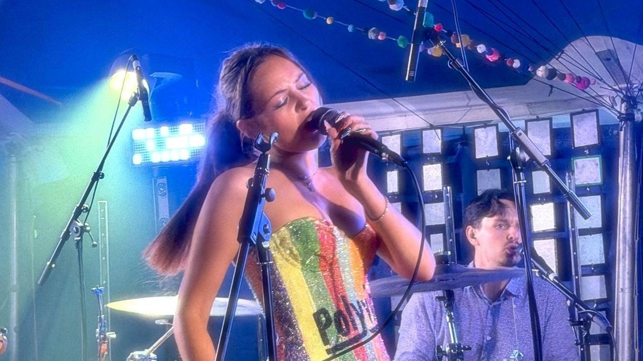 Members of the band Polyvinyl performing on stage. The front-woman is singing into a microphone and wearing rainbow-coloured top. The drummer behind her is also singing into a microphone and is wearing a blue shirt.