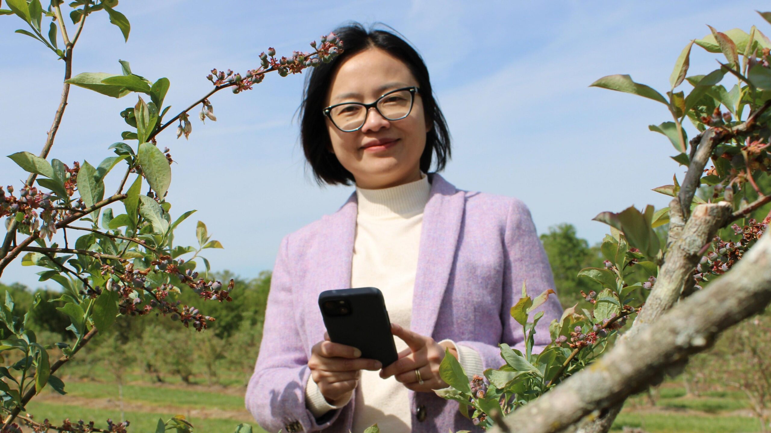Wearing glasses and a lavender-coloured jacket, Jing Zhang stands in an orchard holding her phone.