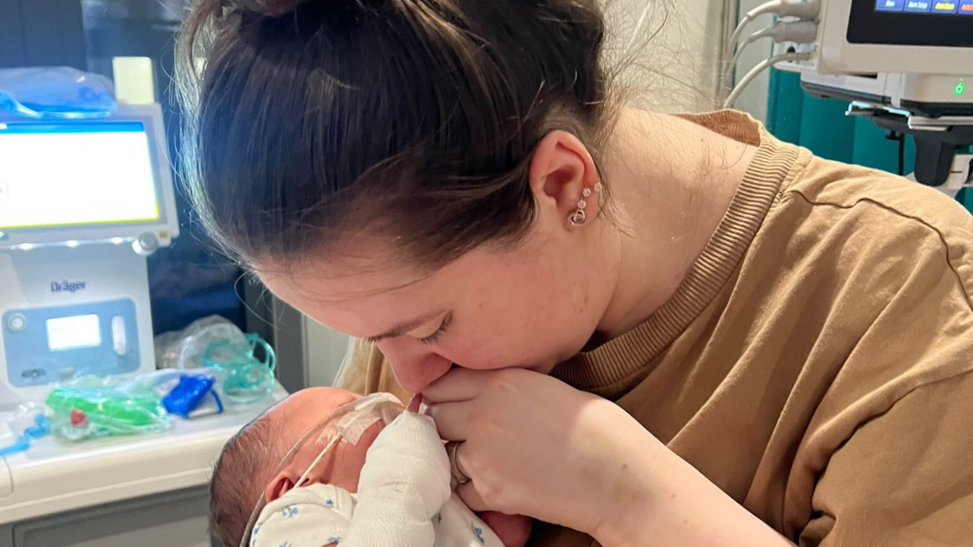 Mum of critically ill baby denied hospital food as she wasn’t breastfeeding Sophieholding her newborn daughter Florence. Tubes can be seen attached to Florence's face. Her left arm is bandaged. Sophie has brown hair which is tied back. She wears a beige t-shirt. Her eyes are closed and she is not looking at the camera.