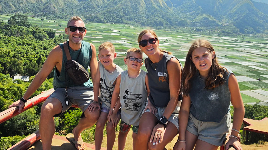 Family of five sitting on a wooden fenced ledge with rice padis in the background and green hills behind . Tom is 38, white, short fair hair. Morgan and George have short fair hair. Dani has long fair hair in a ponytail and Olivia has loose long dark blonde hair. All five wear shorts and sleeveless tshirts in varying shades of grey. Dani and Tom wear sunglasses. They all smile at the camera.