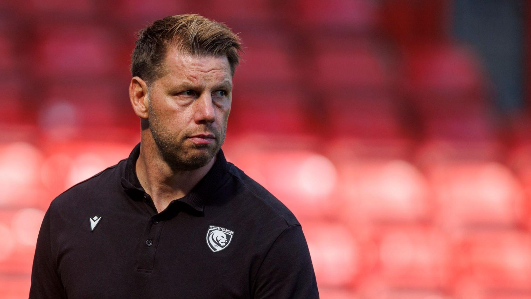 A close-up of Brett Deacon looking on wearing a black polo top with a white Gloucester Rugby badge