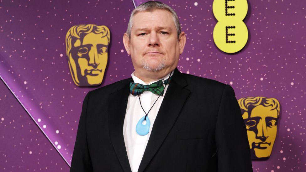 John Davidson is wearing a black suit jacket, white shirt and green tartan bow tie at the Bafta Film Awards in London. He is also wearing a blue pendant round his neck and is standing in front of a screen that features images of the Bafta "mask"