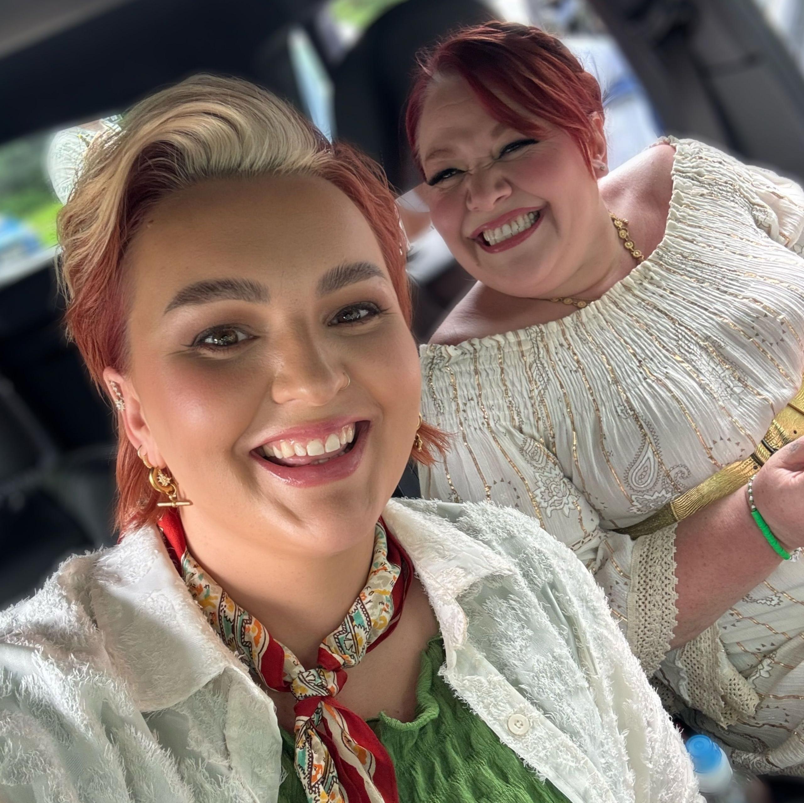 Two women smile at the camera. They both wear white and are sat in a car.