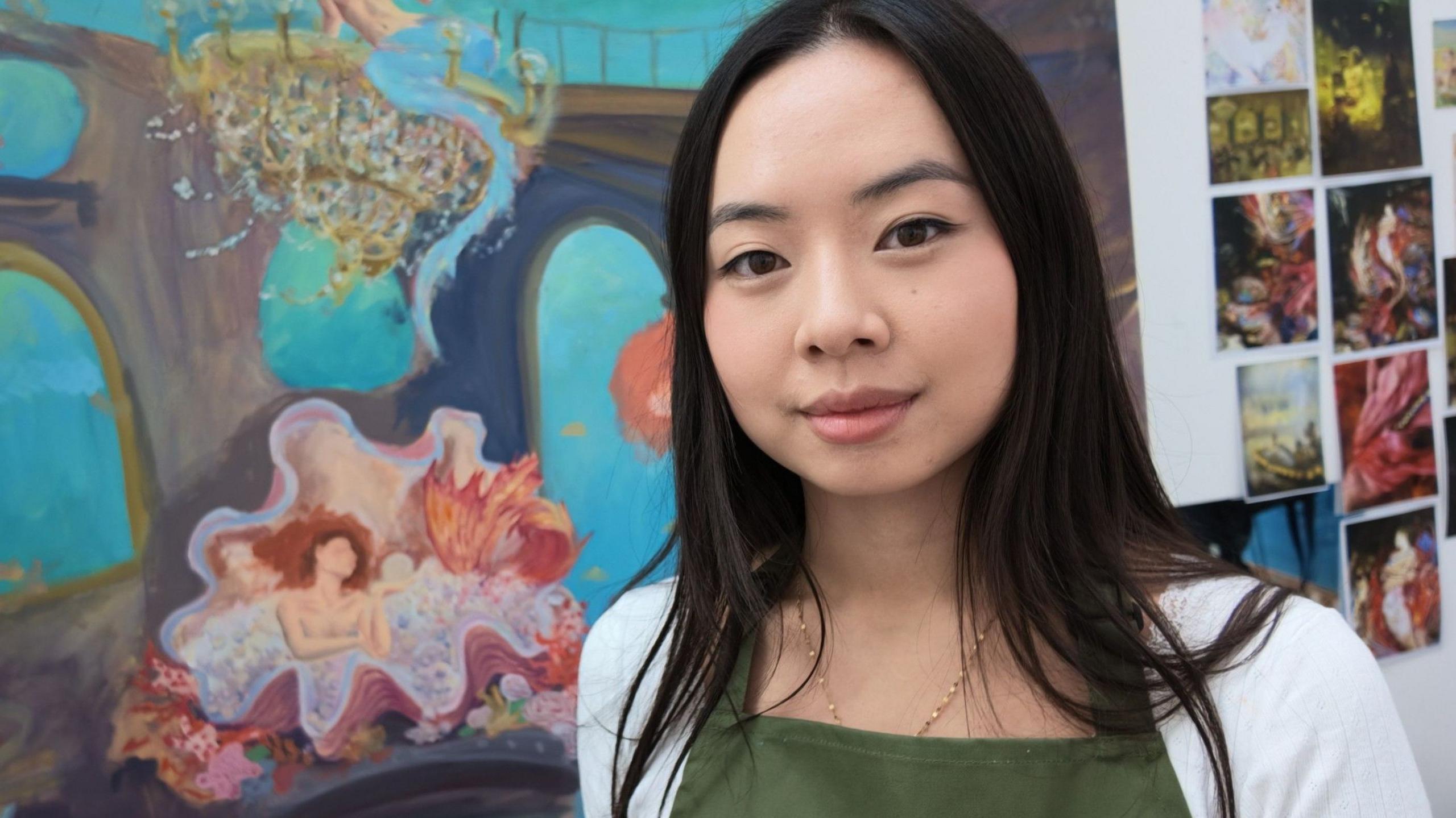 Ivy Deng seen with long, dark hair wearing a white top and green apron looks towards the camera, standing in front of colourful paintings and a board featuring artistic study photos.