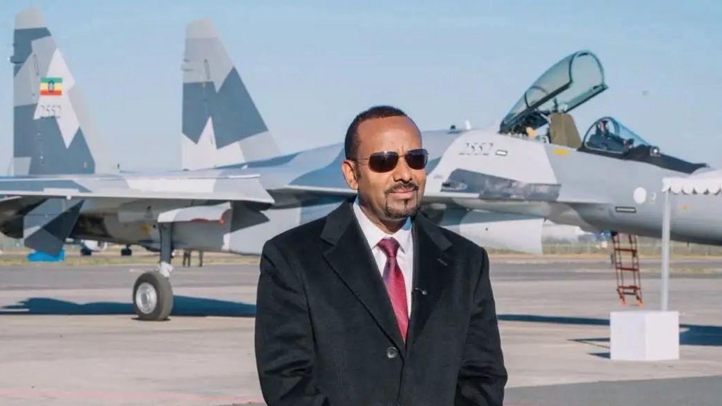 Abiy Ahmed, in a black suit and red tie  standing on tarmac in front of a fighter jet