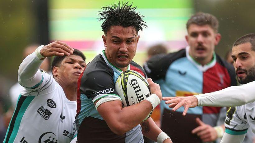 Marcus Smith (centre) has the ball in his right hand/arm as two Vannes players reach out with their hands to try to stop him
