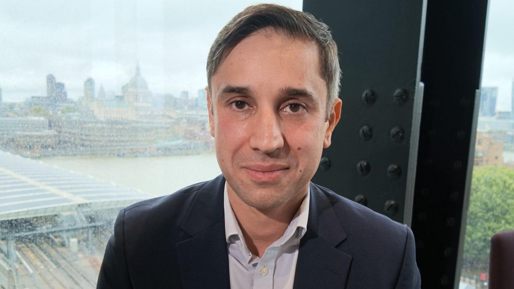 Nick Buckland, a man in a dark suit jacket and open-collared white shirt, looking directly at the camera with a blurred London cityscape visible through the window behind him.