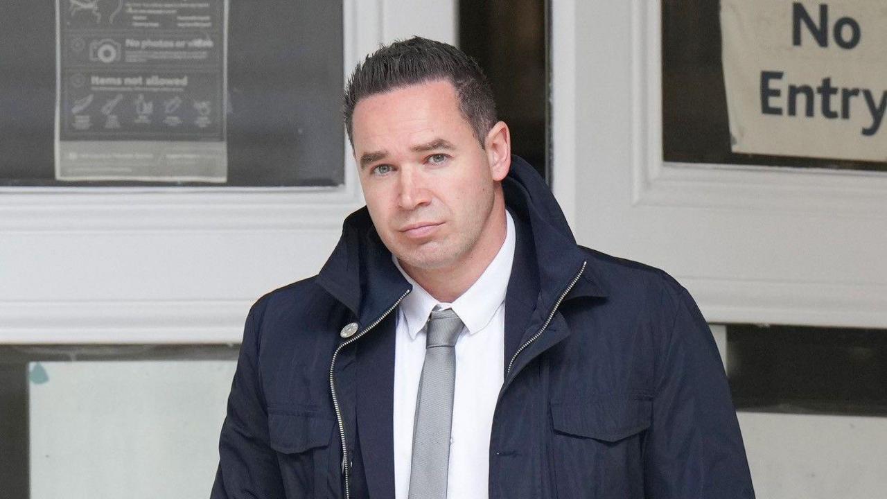 A man with short black hair wearing a large dark blue coat. He is looking into and walking towards the camera.