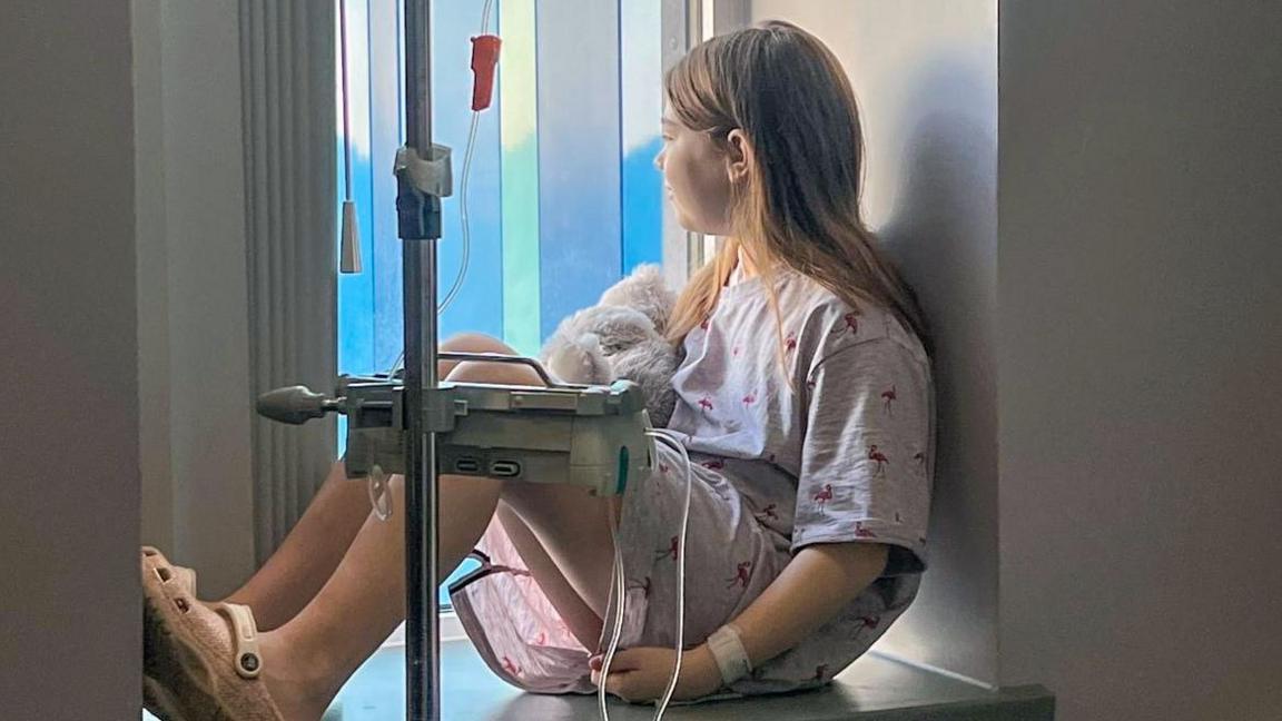 A little girl of around 13 years old with long straight brown hair tucked behind her ears sits half silhouetted in a hospital window attached to a drip with a teddy bear in her lap. She is looking away from the camera and out of the window.