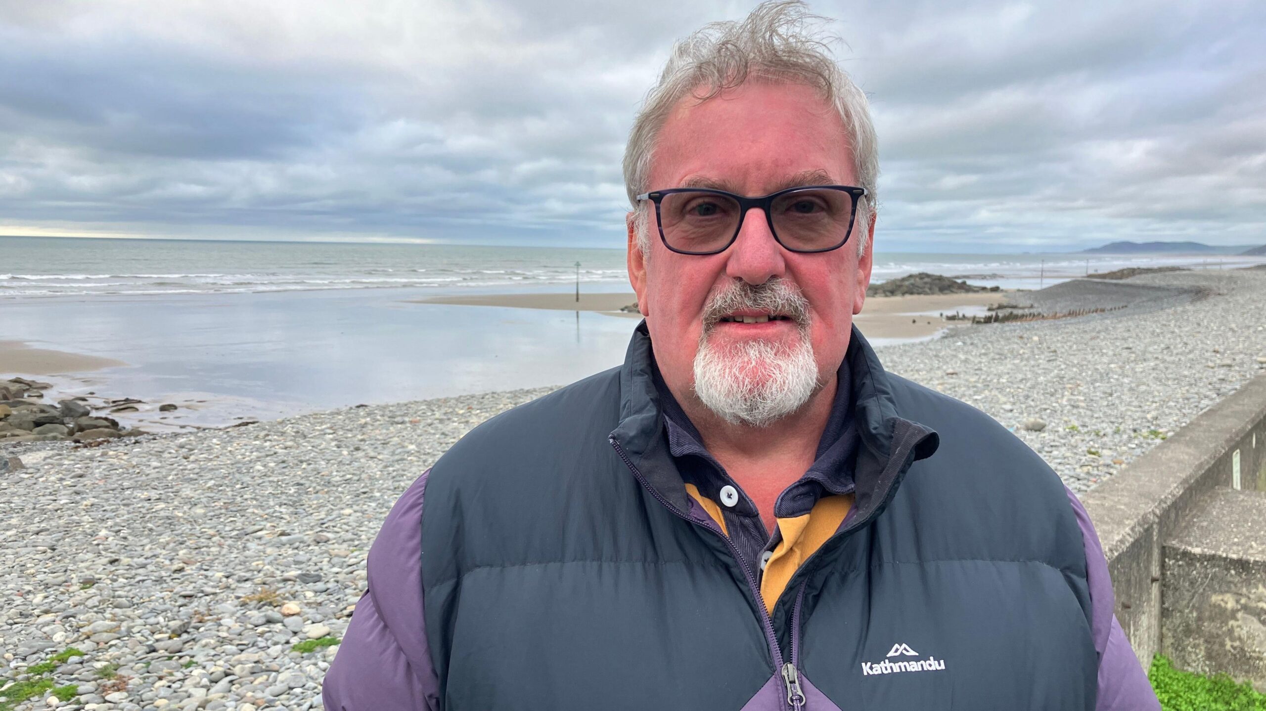 A man with grey hair and a goatee beard and moustache, wearing dark framed glasses and a navy blue and purple body warmer. He's looking straight at the camera and is standing in front of a pebble beach with the sea and sandbanks behond him in the middle distance