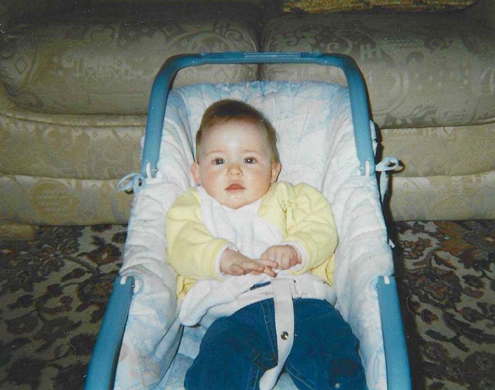 A photo of Courtney-Grace as a baby in a baby seat with a couch in the background