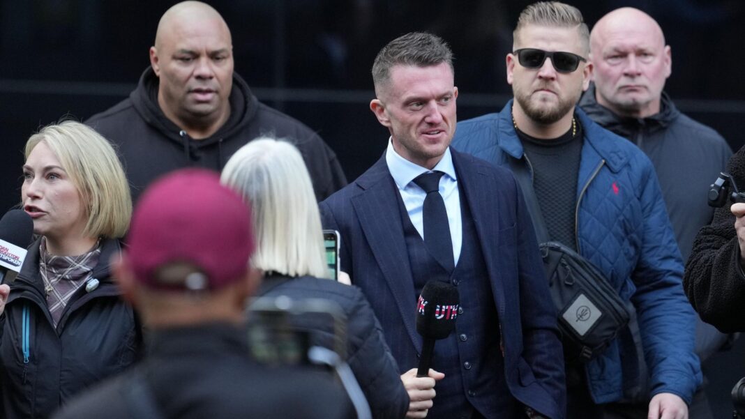 Trial hears Robinson ‘intimidated’ Daily Mail reporters Tommy Robinson (centre) arrives at Southwark Crown Court. He holds a black microphone with the letters UTK on the cover, coloured in the British flag colours. A group of others accompany him. He wears a navy blue suit with a white shirt and blue tie.