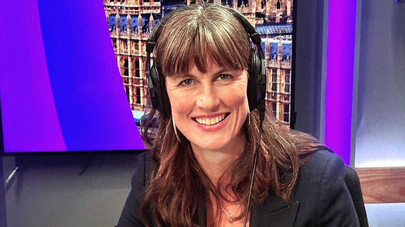 Green Party co-deputy leader Rachel Millward. She is sitting in a BBC studio smiling at the camera. She has long brown hair with a fringe, and is wearing headphones and a black jacket. Behind her are purple-lit walls and an image of the Houses of Parliament.