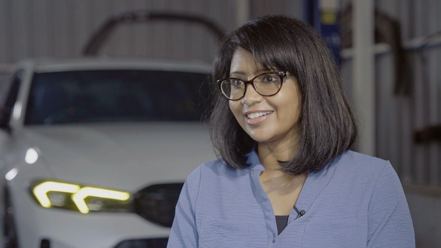 Indian drag racer and IT professional Uvashri Kannadasan Balajeyanthi, wearing glasses and a blue shirt and smiling, in front of a white car inside a garage.