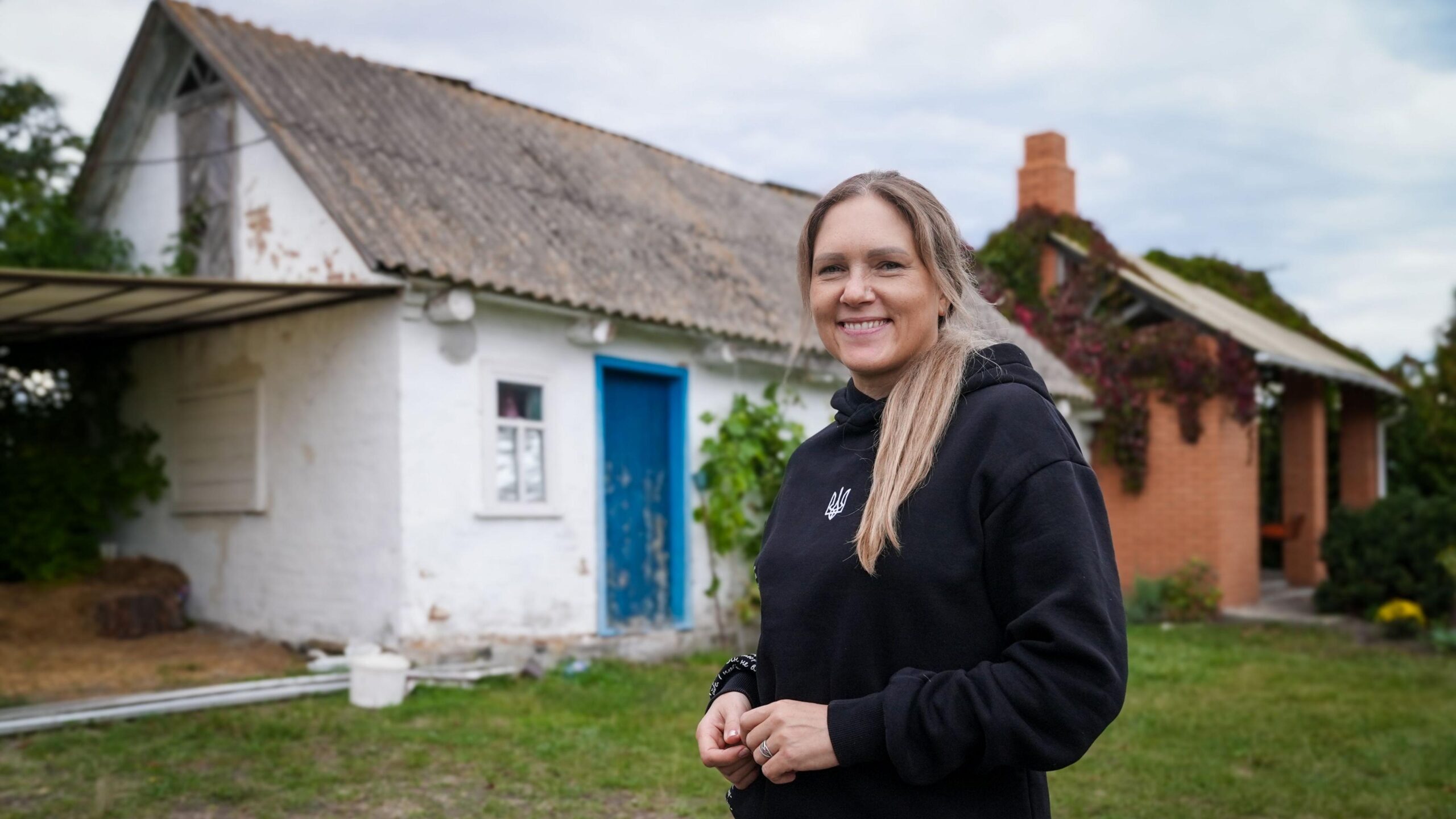 A woman with long hair wearing a black hoodie stands in front of a modest white building with a blue door.