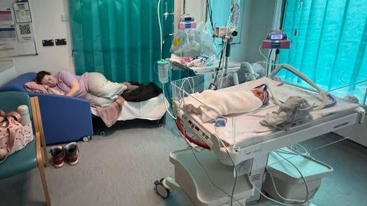 Inside the paediatric ward at Grange University Hospital in Cwmbran. Sophie Price is a young woman who seen lying on her side on top of a sofa bed. Her daughter Florence is inside a hospital baby crib. A breathing tube can be seen over her nose. She is wrapped in a white blanket.