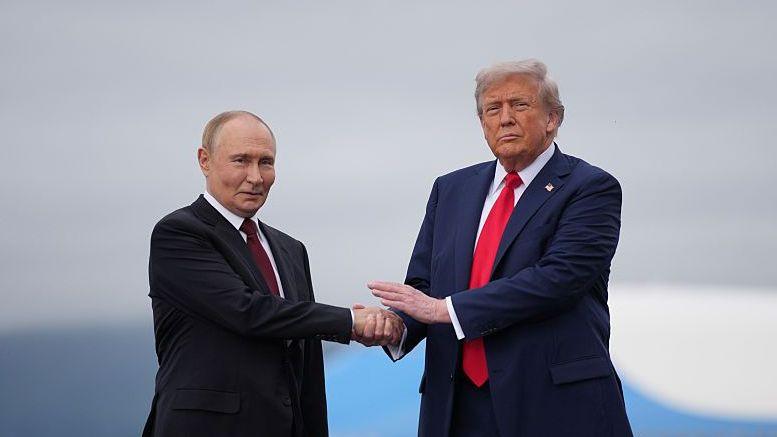 Two men shake hands as they pose for photographs. The shorter man, to the left, wears a black suit jacket and maroon tie, while the taller man, to the right, wears a dark blue suit jacket and red tie.