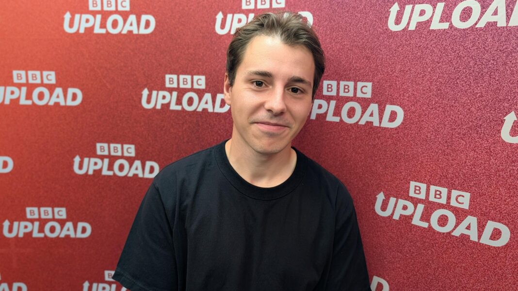 Benjamin has long brown hair which is swept back and he is smiling. He is wearing a black T-shirt and is standing in front of a red glittery backdrop that has white text on it reading 