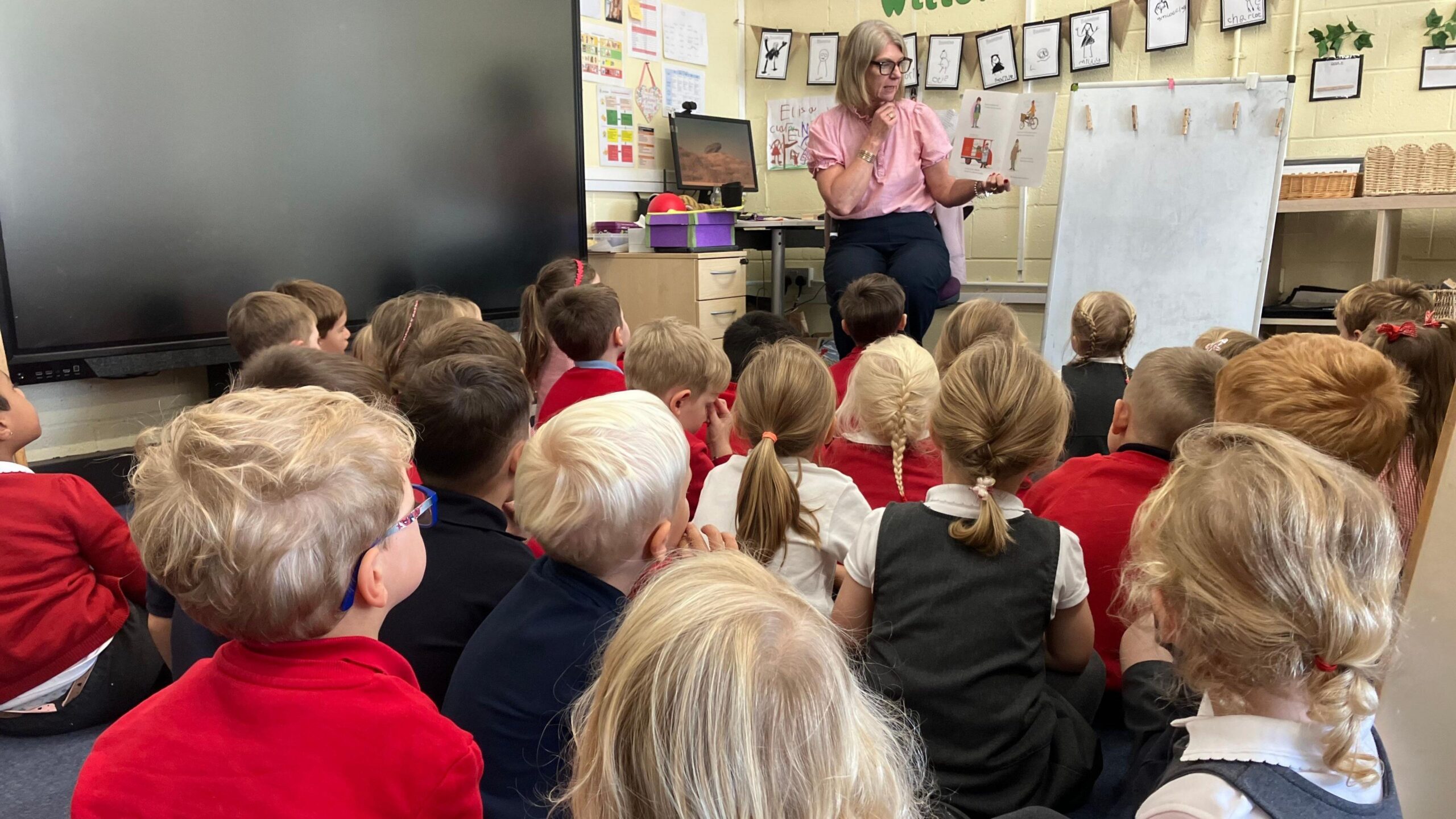 Teacher seated at the front of a classroom reading from a picture book to a large group of young children sitting on the floor. A whiteboard, display screen and children's artwork are visible around the room.