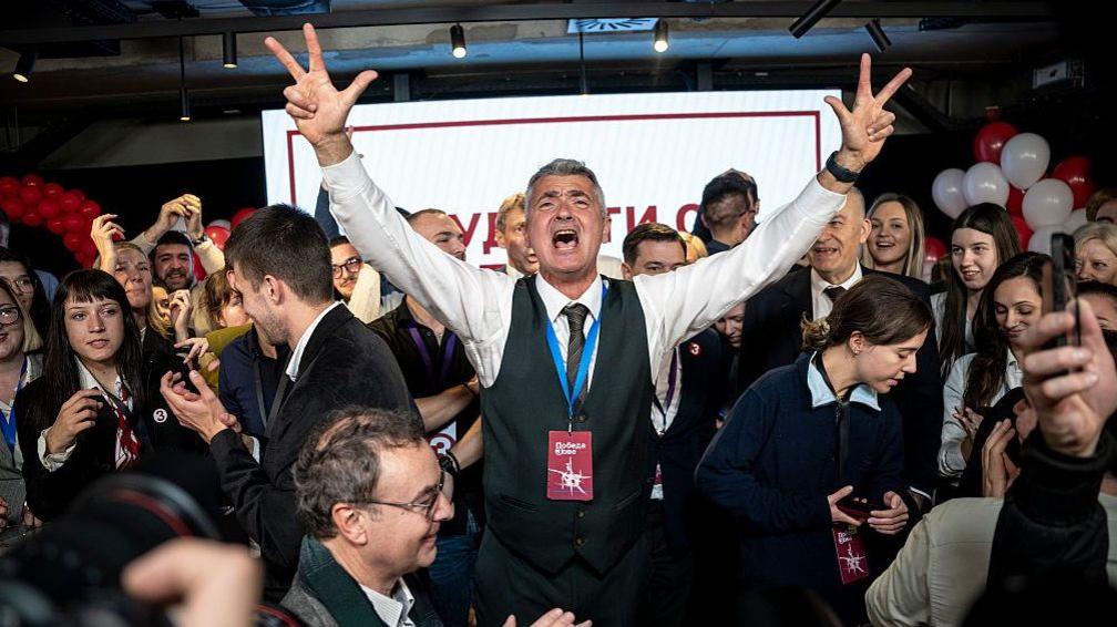 A man in a waistcoat and grey hair celebrates