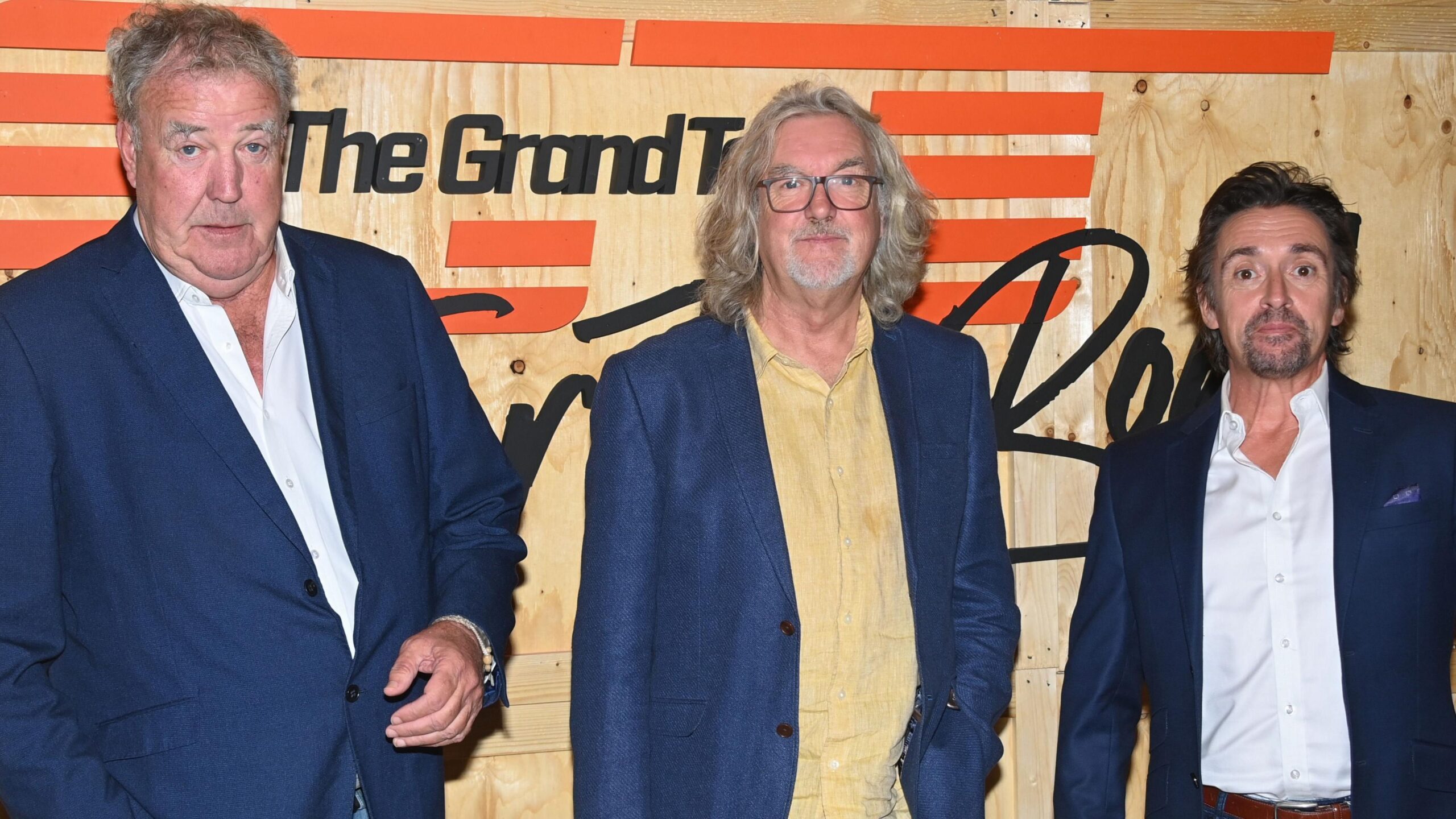 L-R Jeremy Clarkson, James May and Richard Hammond. They are wearing blue suits and standing in front of promotional sign for The Grand Tour TV programme