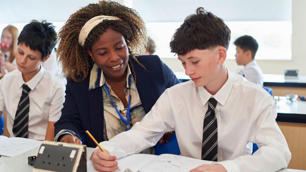 Ofsted faces fresh calls to halt graded school inspections in England Teacher supports a student during a classroom lesson, looking over written work at a science lab bench. Other students in school uniform work in the background, with educational equipment and diagrams visible on desks.