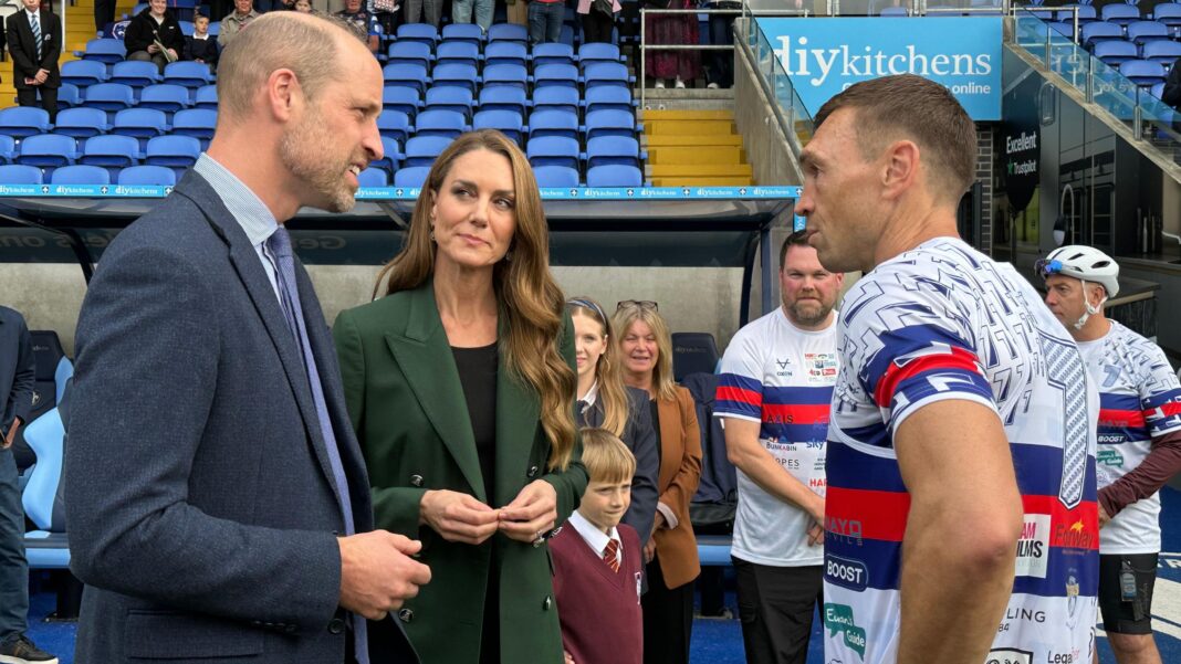 Sinfield completes fundraising challenge of seven ultramarathons in a week Three people stand beside a stadium dugout during a sporting event. A person in a navy suit and tie speaks with a person wearing a dark green double-breasted blazer, while a rugby player in a white jersey with blue and red horizontal stripes faces them. Behind the group, spectators and team members stand near the bench area, with rows of empty blue seats rising in the background. Visible signage includes “diykitchens” on a board above the tunnel entrance. Another player in matching kit and a cyclist wearing a helmet can be seen to the right of the image.
