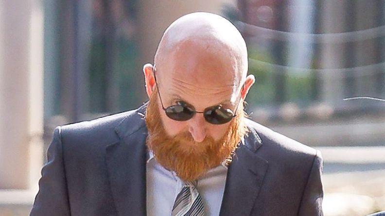 Neil Lewis wearing a suit. He is bald, wearing sunglasses and has a long ginger beard.