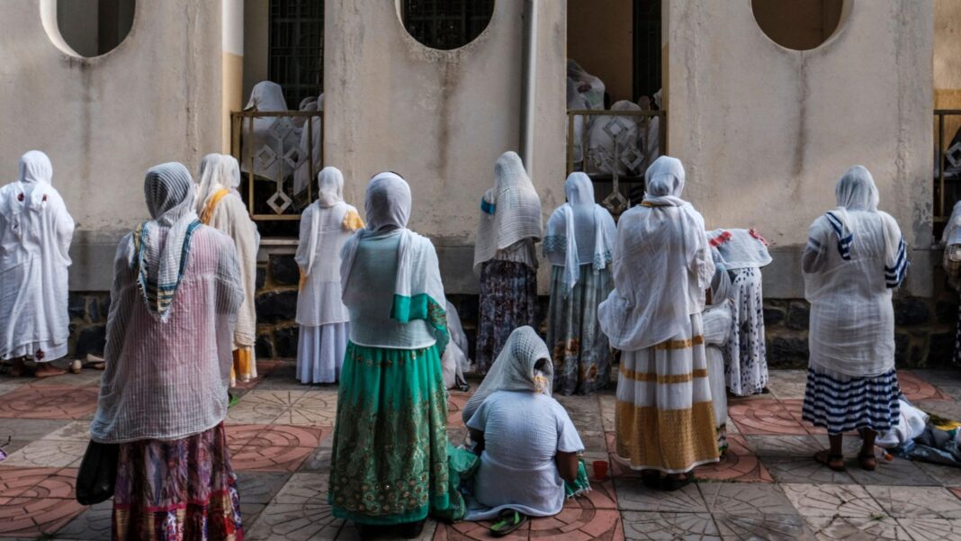 More than 60 Christians killed in Ethiopia ‘massacre’, church official says Women wearing colourful traditional Ethiopian dresses in green, red and gold, with long headscarves, pray outside a church. Some stand while others kneel.