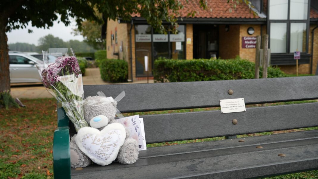 Woman found dead in home named as GP A wooden bench with green arms is shown in the foreground with a grey teddy bear, a bunch of flowers and a small card of condolence placed on it. Behind it is a medical centre, parked cars, trees and other greenery.
