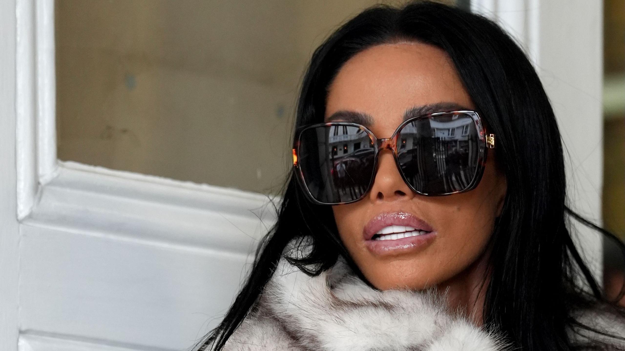 A close up of Katie Price arriving at Lewes Crown Court, she is wearing large dark sunglasses and looking over her shoulder. She has dark hair and is wearing a pale fur jacket, the white wooden doors of the building can be seen behind her.