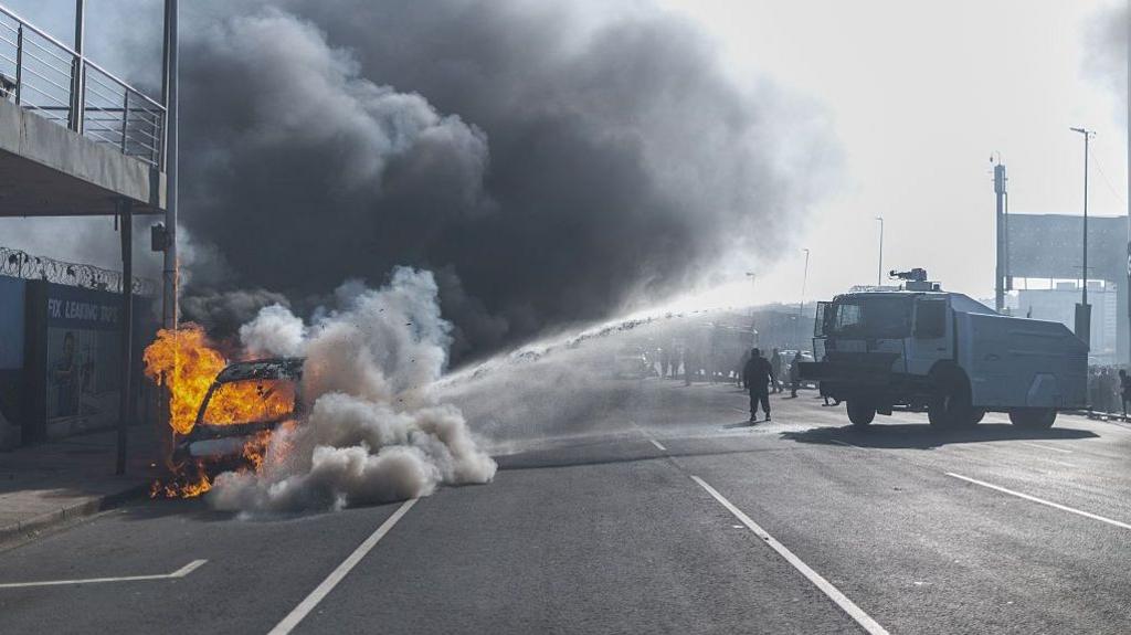 A vehicle burns on the side of a road as thick black smoke fills the air, while a police water-cannon truck sprays water towards the scene