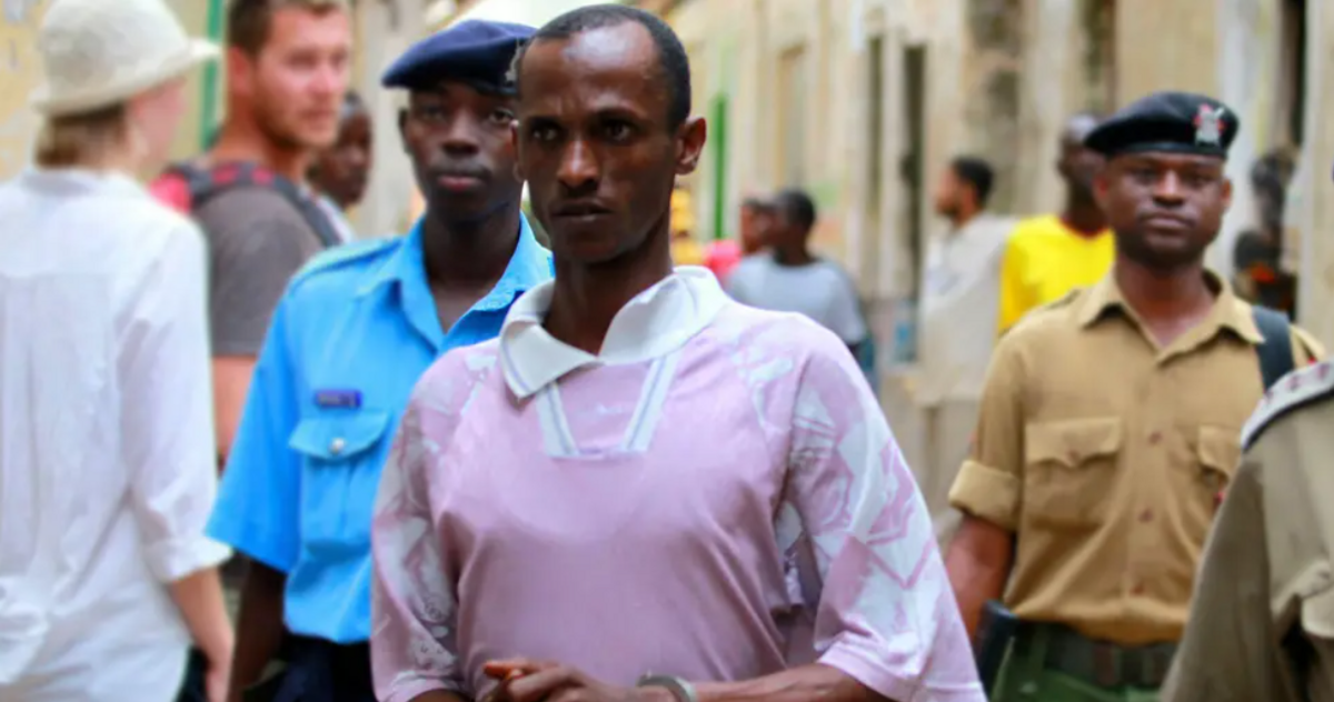 Ali Kololo escorted by police in handcuffs wearing a pink t-shirt with a white collar