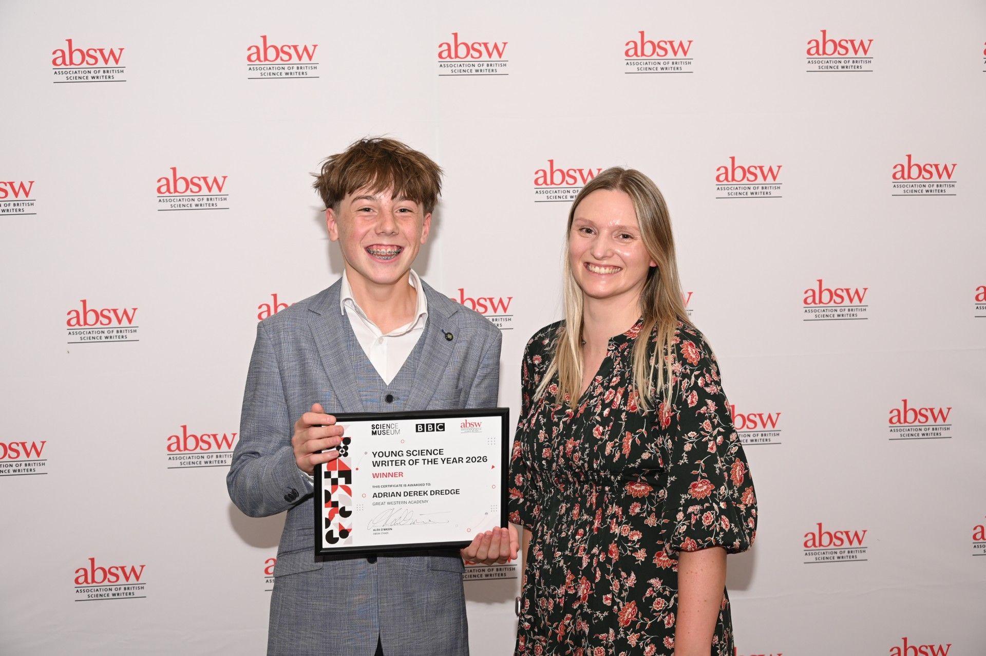 Adrian Dredge holds a certificate annoucing him winner of the Young Science Writer of the Year for 2026. He is wearing a grey suit and white shirt, and stands beside his teacher, Charlotte Whittenbury, from Great Western Academy in Swindon.