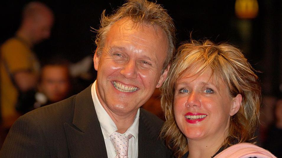 Sarah Fisher, with her blonde hair tousled in a long crop and wearing a pink shawl, with Anthony Head who is wearing a black jacket, white shirt and tie. The pair have big smiles for photographers.