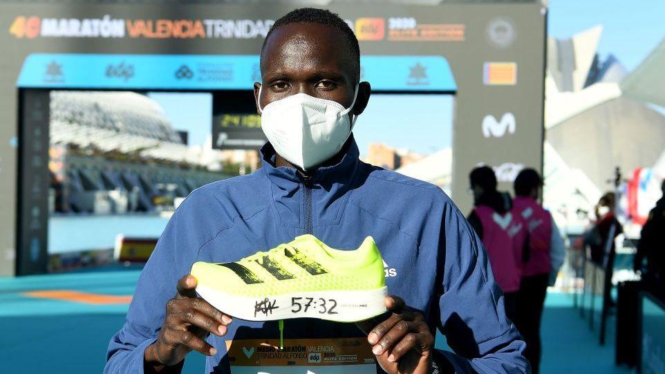 A medium close-up photograph features Kenyan long-distance runner Kibiwott Kandie posing with a running shoe after setting a record. He faces the camera wearing a blue athletic jacket, a white face mask, with both hands, holding up bright neon-yellow Adidas running shoe at chest level, which has the time "57:32" written in black marker across its thick white midsole.